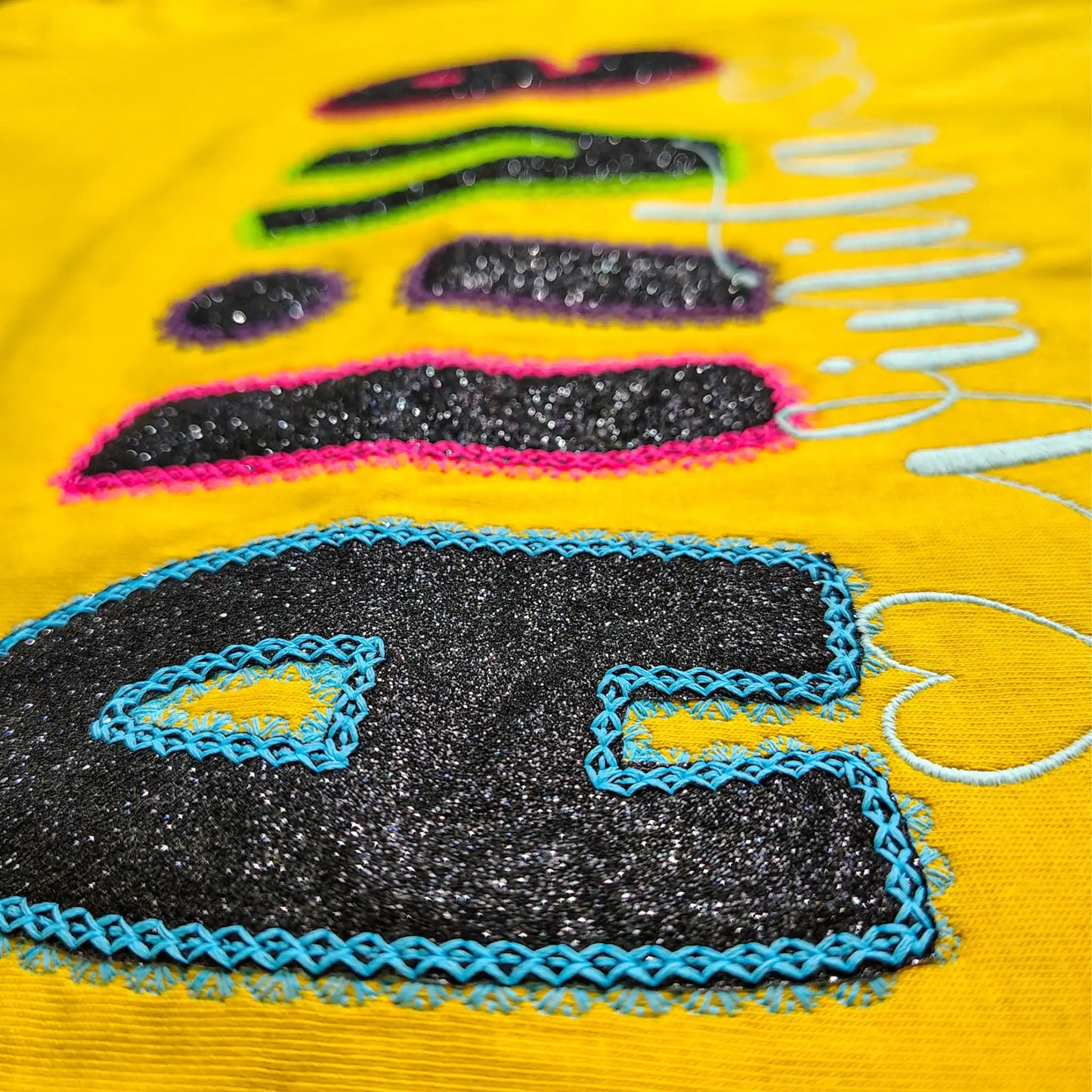 Macro shot of glitter appliqué letter detail — multicolour fabric fill with neon embroidery outline on yellow cotton, by Embroidist.