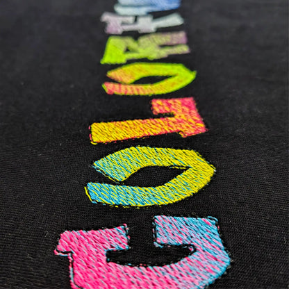 Macro shot of multicolour COLORFUL embroidery on black cotton — rainbow satin stitch lettering with green leaf prop, by Embroidist.