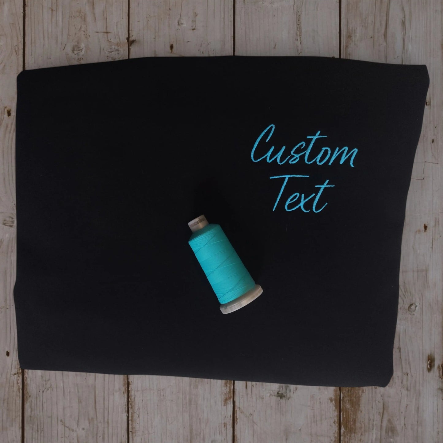 Black t-shirt with teal cursive custom text embroidery and matching thread spool — left chest placement by Embroidist