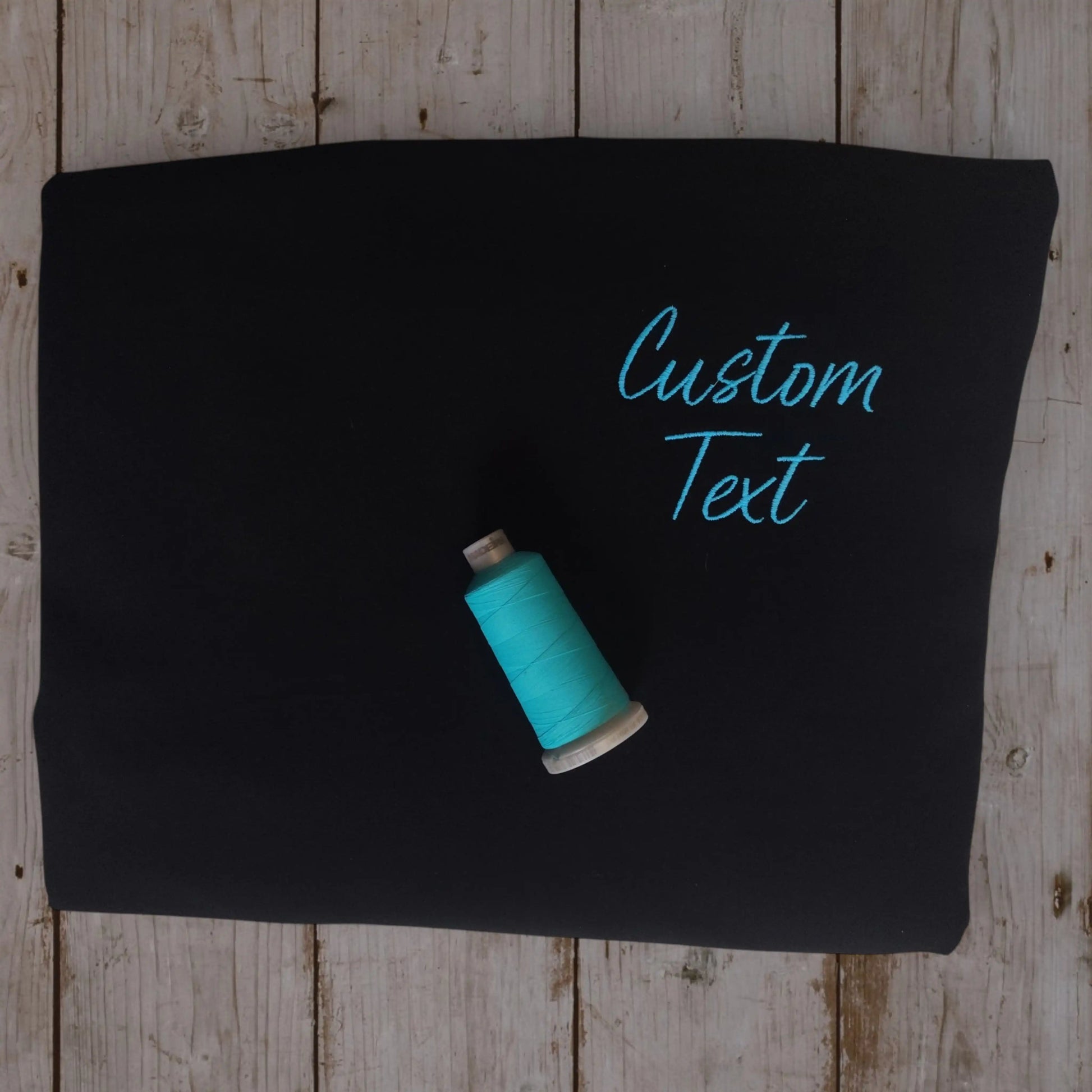 Black t-shirt with teal cursive custom text embroidery and matching thread spool — left chest placement by Embroidist