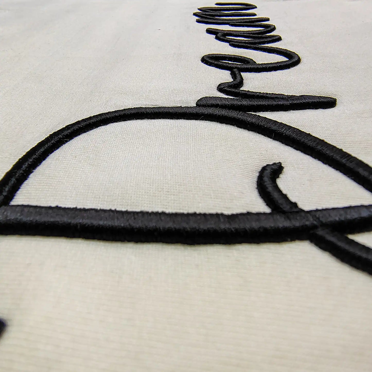Extreme close-up of 3D puff embroidery thread detail on cream fabric — black raised cursive stitch showing depth, shadow and texture of Dream tee, by Embroidist