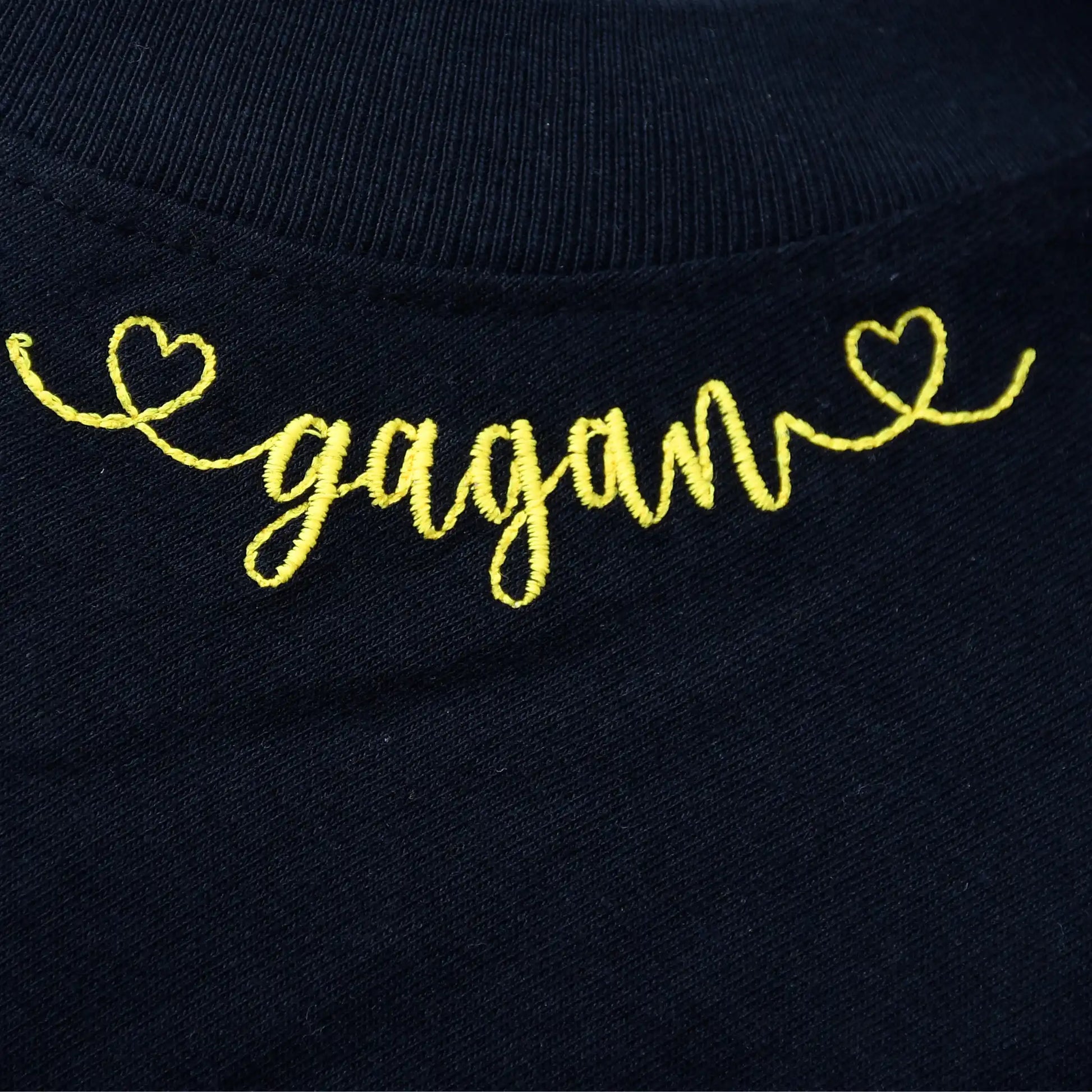 Personalised name "Gagan" embroidered in yellow cursive script with heart motifs at neckline on black t-shirt — custom name embroidery detail by Embroidist.