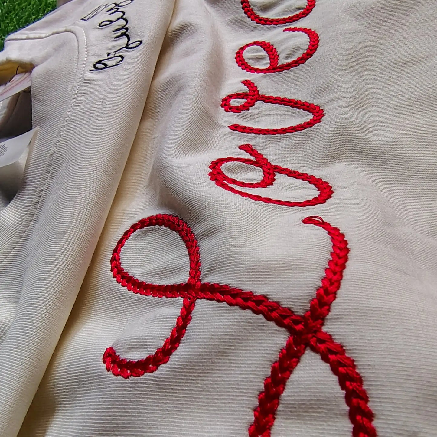 Macro shot of red chain stitch "Loved" embroidery and date of birth on neckline— individual stitch loops visible on cream cotton fabric, by Embroidist.