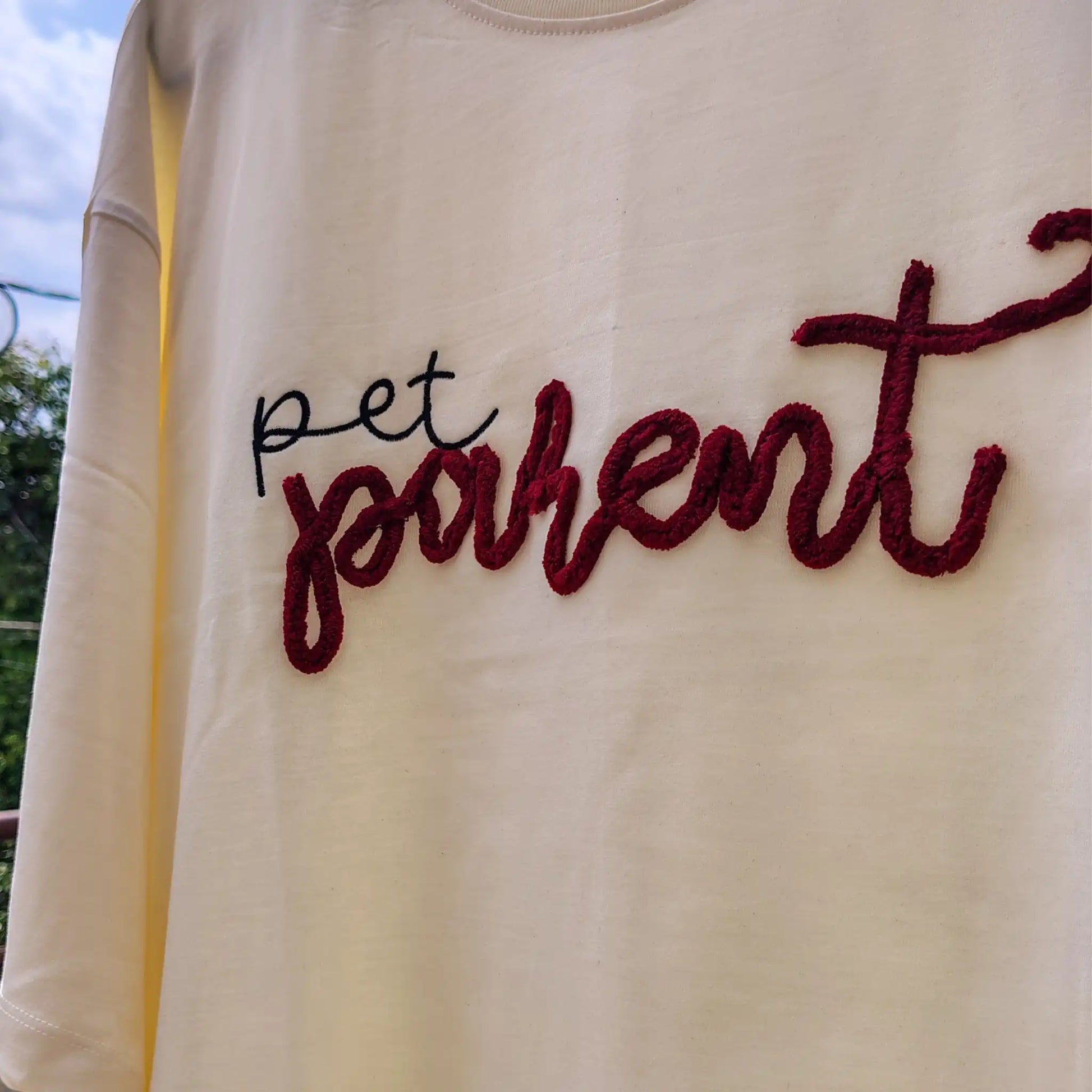 Flat lay of cream t-shirt with "pet parent" red and black chenille script — full front view showing left chest embroidery placement, by Embroidist.