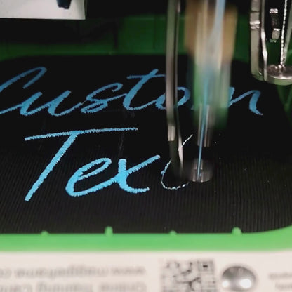 Embroidery machine stitching teal cursive custom text on black fabric — live production at Embroidist