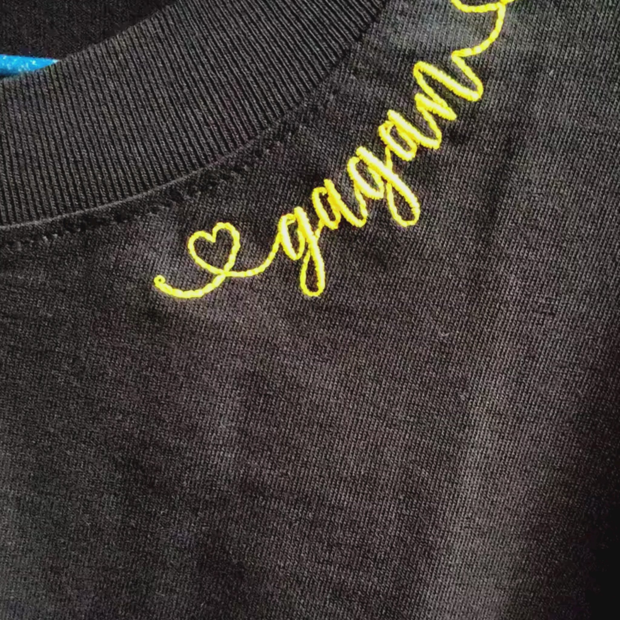 Video of personalised "♡gagan" name embroidery in gold cursive thread at neckline — close-up detail showing heart motif and script stitch on black fabric, by Embroidist.