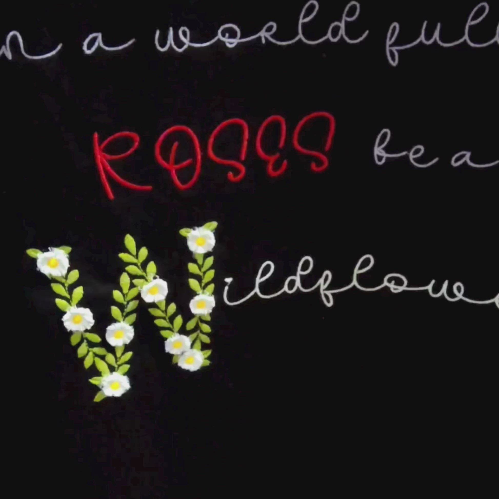 Close-up video of Wildflower floral embroidery detail — white daisy blooms, green leaves and multicolour lettering on black fabric, by Embroidist.