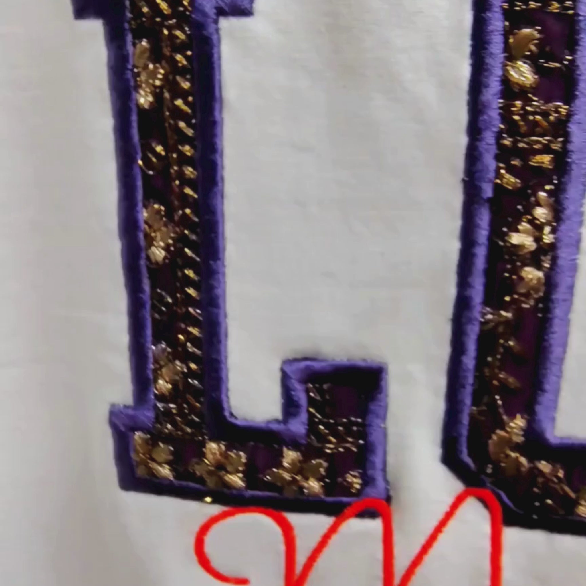 Close-up video of Zari appliqué LOVE lettering detail — gold embroidered fill with purple border on cream fabric, by Embroidist.