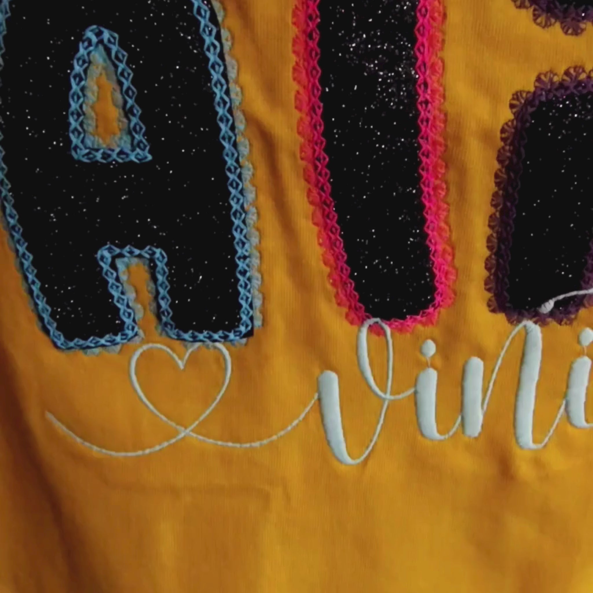 Close-up video of ALIVE glitter appliqué embroidery detail — multicolour letters with neon outline stitch on yellow fabric, by Embroidist.