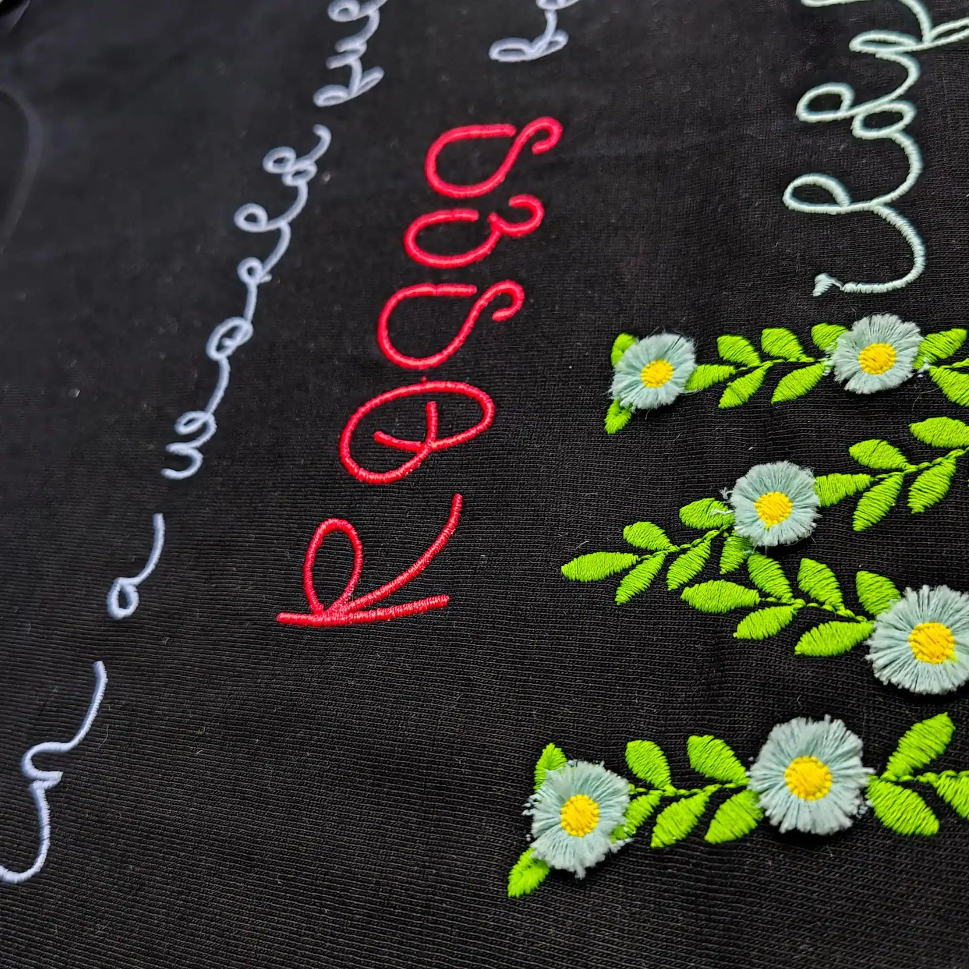 Macro shot of white daisy and green leaf embroidery detail — floral chain stitch on black cotton fabric, by Embroidist.