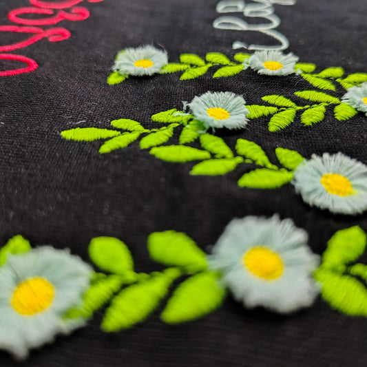 Flat lay of black t-shirt with "in a world full of roses be a Wildflower" embroidery — multicolour floral script with botanical details, by Embroidist.