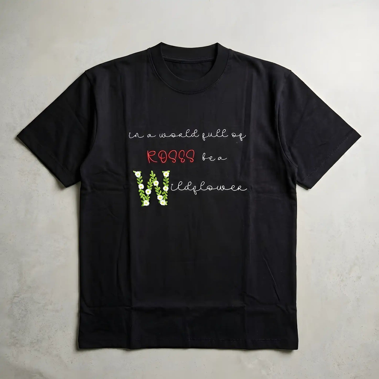 Wildflower floral embroidery t-shirt flatlay — "in a world full of roses be a wildflower" satin stitch floral lettering on black oversized tee, machine embroidered in India by Embroidist