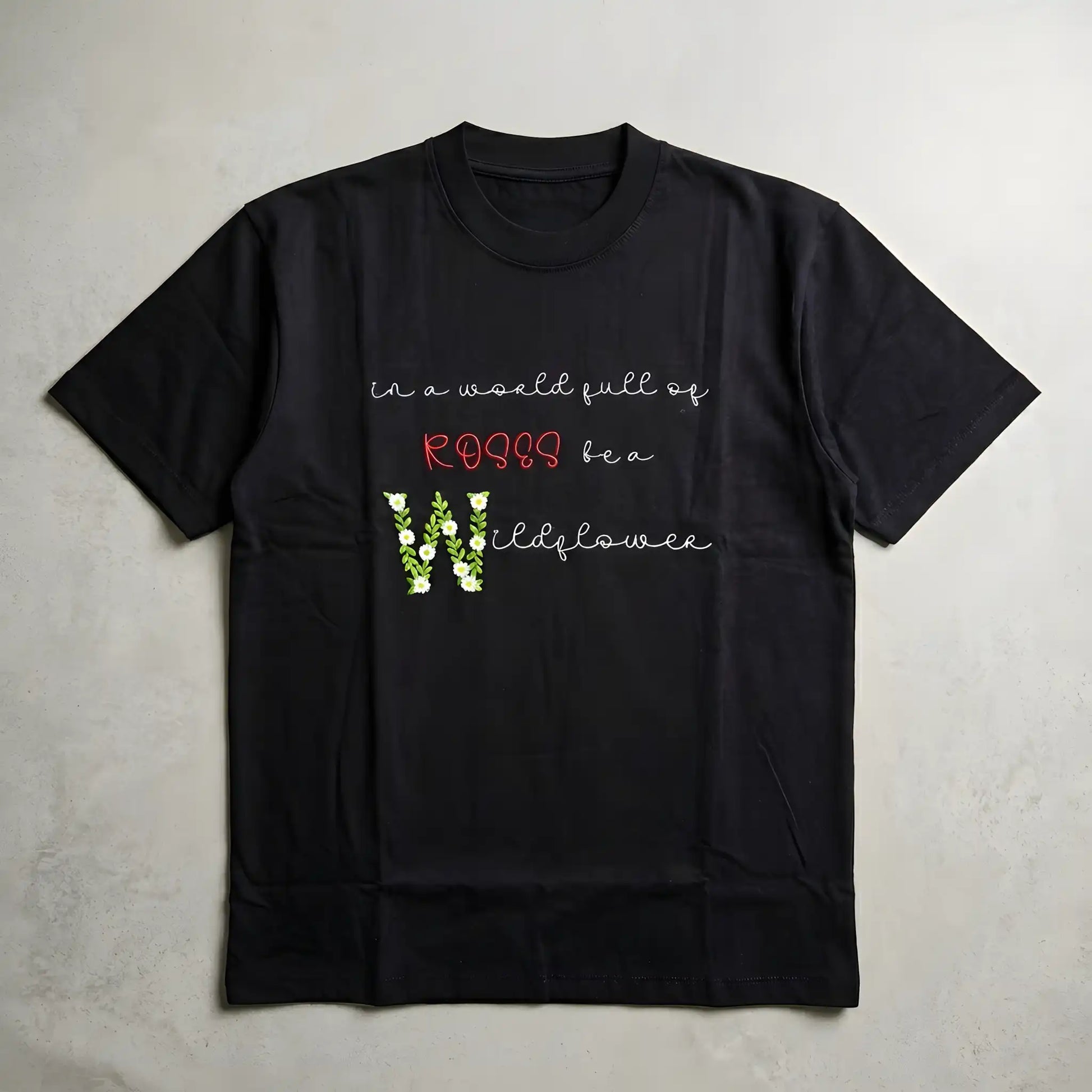 Wildflower floral embroidery t-shirt flatlay — "in a world full of roses be a wildflower" satin stitch floral lettering on black oversized tee, machine embroidered in India by Embroidist