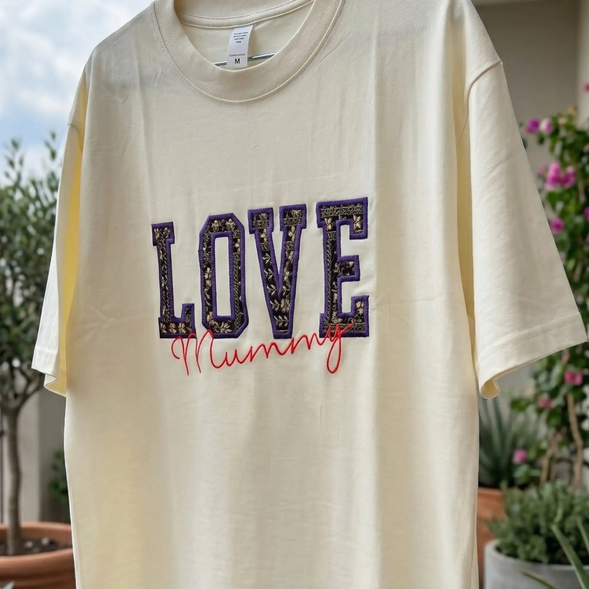 Flat lay of cream LOVE Zari embroidered t-shirt — full front view showing appliqué typography and cursive name personalisation, by Embroidist.